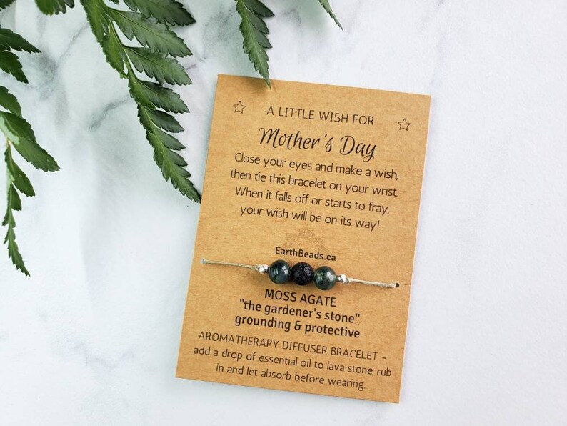 May include: A brown card with the text "A Little Wish For Mother's Day" and a green and black moss agate bracelet with a lava bead. The card says to close your eyes and make a wish, then tie the bracelet on your wrist. When it falls off or starts to fray, your wish will be on its way! EarthBeads.ca. The bracelet is described as a Moss Agate "the gardener's stone" grounding & protective aromatherapy diffuser bracelet. Add a drop of essential oil to lava stone, rub in and let absorb before wearing.