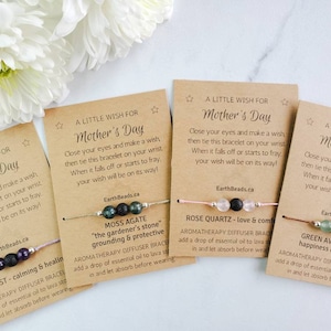 May include: Four Mother's Day bracelets with cards. Each bracelet has a different type of stone bead and a lava bead. The cards have a message about making a wish and tying the bracelet on your wrist. The stones are amethyst, moss agate, rose quartz, and green aventurine.