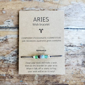 Peut inclure: Une carte brune avec le texte "ARIES Wish bracelet" et le symbole astrologique du Bélier. La carte comprend également le texte "CONFIDENT/PASSIONATE/COMPETITIVE jade, bloodstone, aquamarine, green aventurine" et "EarthBeads.ca". Un bracelet en cordon noir avec des perles vertes, brunes et bleues est présenté sur la carte.