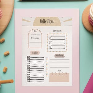 Op de afbeelding: Een roze "Daily Flow" planner met secties voor datum, affirmaties, schema, topprioriteiten, brainstorm en notities. De planner is omgeven door kleurpotloden, een cupcake, bloemen en een kopje thee.