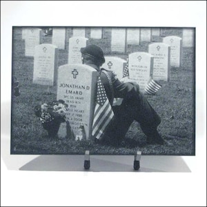 May include: A black and white photo of a soldier in uniform kneeling in front of a grave marker. The grave marker is inscribed with the name "Jonathan D. Emard" and the words "SPC US Army Iraq KIA BSM Purple Heart May 37 1985 SN 4 2005  Forgotten Late". The soldier is holding an American flag.