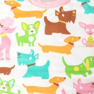 May include: A white baby bib with a pink trim, featuring a repeating pattern of cartoon dogs in various colours. The dogs include dachshunds, poodles, and terriers, with collars and bows.