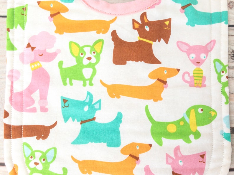 May include: A white baby bib with a pink trim, featuring a repeating pattern of cartoon dogs in various colours. The dogs include dachshunds, poodles, and terriers, with collars and bows.