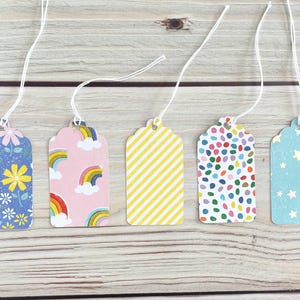 May include: Five decorative gift tags with white string. The tags feature various patterns: floral, rainbows, yellow stripes, colorful confetti, and stars. Each tag is a different color, including blue, pink, and yellow.