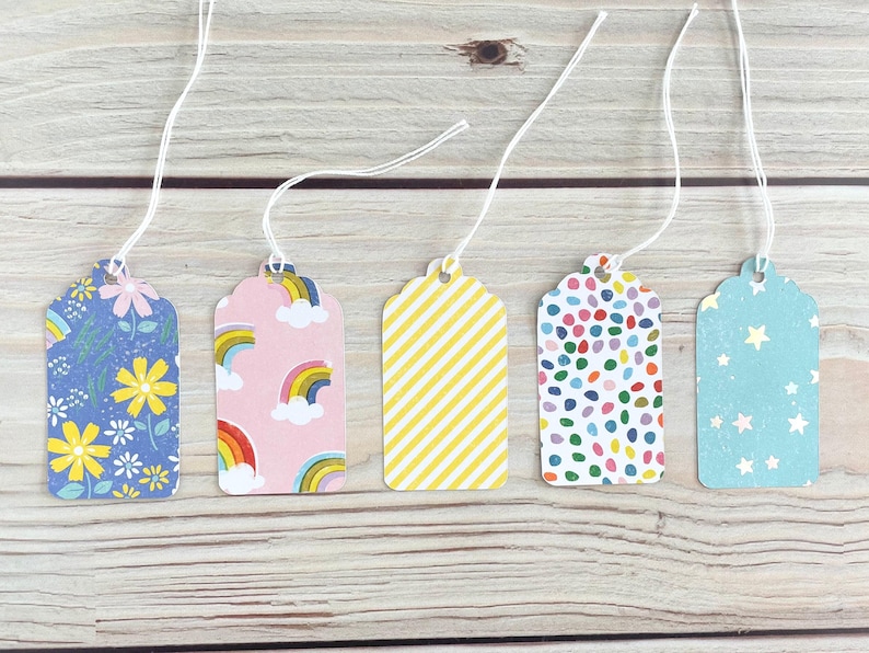 May include: Five decorative gift tags with white string. The tags feature various patterns: floral, rainbows, yellow stripes, colorful confetti, and stars. Each tag is a different color, including blue, pink, and yellow.