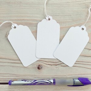 May include: Three white gift tags with scalloped edges and string attached. The tags are lying on a wooden surface with a purple pen.