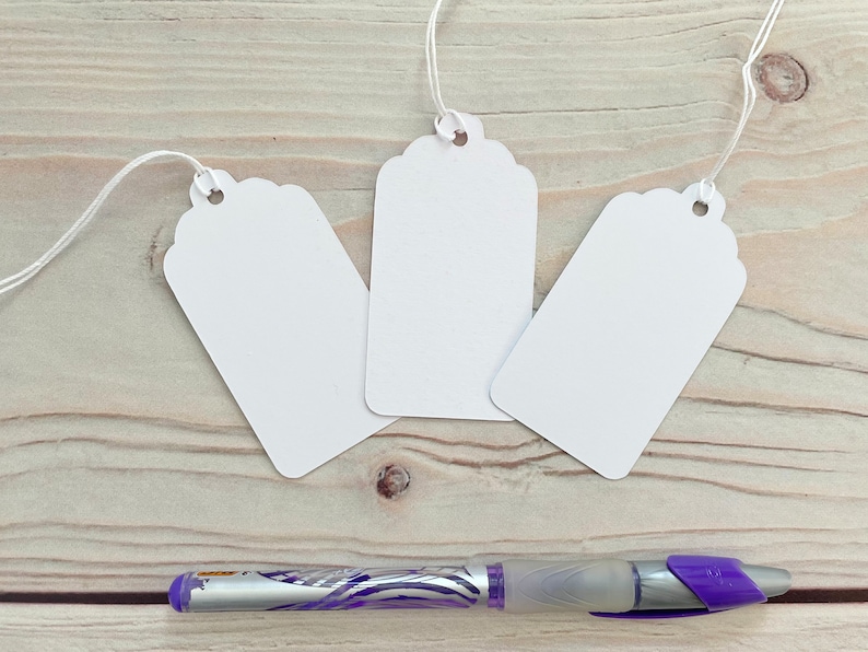 May include: Three white gift tags with scalloped edges and string attached. The tags are lying on a wooden surface with a purple pen.