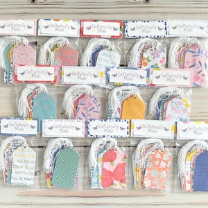 May include: A collection of gift tags in clear plastic packaging. Each tag has a white string and is decorated with various floral and patterned designs in pink, blue, yellow, and orange. The text "Craftastically Made" is printed on each package.