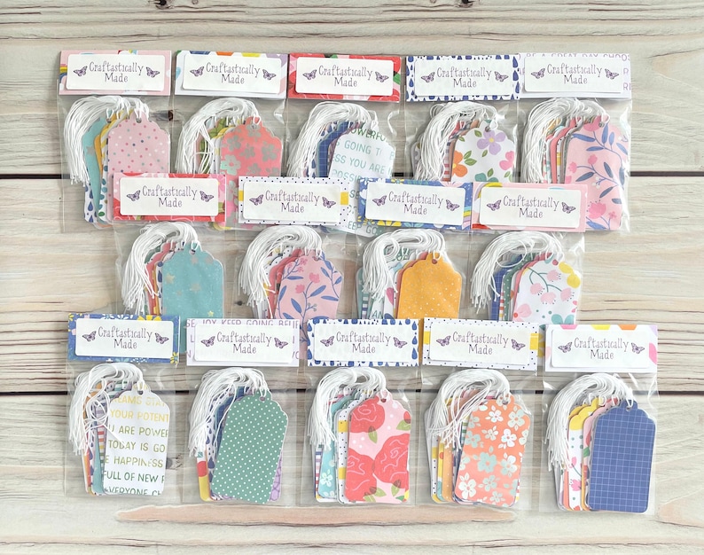 May include: A collection of gift tags in clear plastic packaging. Each tag has a white string and is decorated with various floral and patterned designs in pink, blue, yellow, and orange. The text "Craftastically Made" is printed on each package.