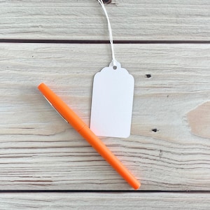 May include: An orange pen and a blank white tag with a scalloped edge are displayed on a light wood grain surface. The pen is angled across the lower left, while the tag hangs from a white string.