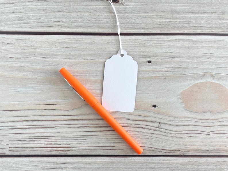 May include: An orange pen and a blank white tag with a scalloped edge are displayed on a light wood grain surface. The pen is angled across the lower left, while the tag hangs from a white string.