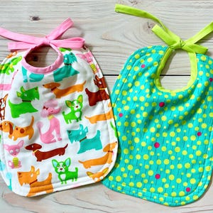 May include: Two baby bibs with tie closures. The left bib is white with a colourful pattern of various dog breeds in green, orange, pink, and brown. The right bib is teal with a pattern of yellow circles and small pink dots, featuring a lime green tie.
