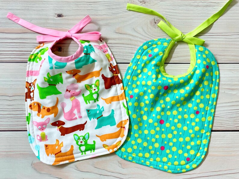 May include: Two baby bibs with tie closures. The left bib is white with a colourful pattern of various dog breeds in green, orange, pink, and brown. The right bib is teal with a pattern of yellow circles and small pink dots, featuring a lime green tie.