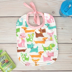 May include: A white bib with a pink trim and a pink tie. The bib has a pattern of colourful dogs on it.