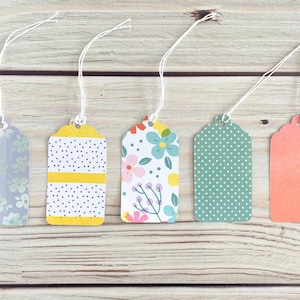 May include: Five decorative gift tags with white string. The tags have various patterns, including floral, polka dots, and stripes, in colors like blue, yellow, and coral. The tags are arranged on a light wood surface.