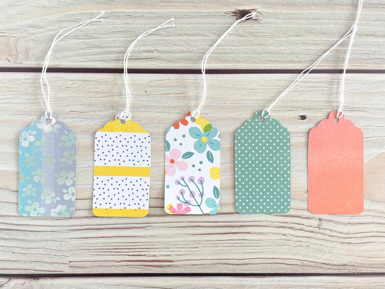 May include: Five decorative gift tags with white string. The tags have various patterns, including floral, polka dots, and stripes, in colors like blue, yellow, and coral. The tags are arranged on a light wood surface.
