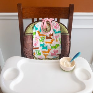 May include: A white high chair tray with a small bowl containing white food and a blue spoon. A bib with a colourful dog pattern and a pink tie sits on the high chair seat cushion, which features brown and green geometric fabric patterns.