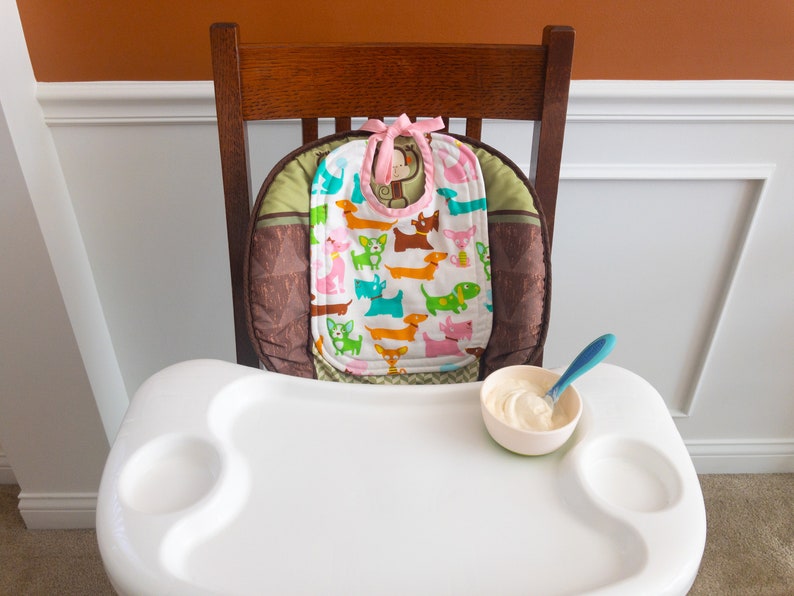 May include: A white high chair tray with a small bowl containing white food and a blue spoon. A bib with a colourful dog pattern and a pink tie sits on the high chair seat cushion, which features brown and green geometric fabric patterns.