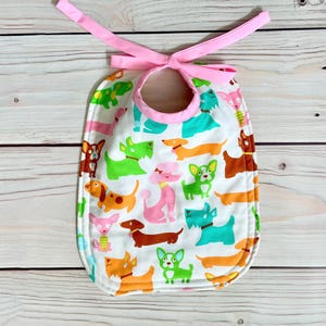 May include: A white baby bib patterned with colourful cartoon dogs in green, orange, pink, and brown. The bib has a pink trim and a matching pink tie closure. Ideal for keeping clothes clean during feeding.
