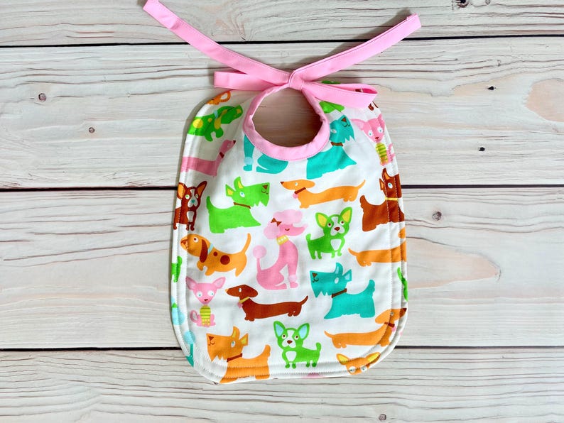 May include: A white baby bib patterned with colourful cartoon dogs in green, orange, pink, and brown. The bib has a pink trim and a matching pink tie closure. Ideal for keeping clothes clean during feeding.