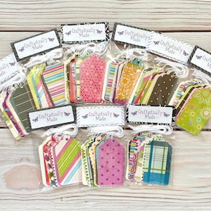 May include: A collection of colorful gift tags in clear plastic bags, arranged on a light wood surface. Each tag features unique patterns and designs, with the text "Craftastically Made" on a label.