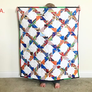 May include: A child holds up a colorful quilt with a white background and a geometric pattern of orange, blue, and red squares. The quilt is made of various fabrics with different patterns and designs.