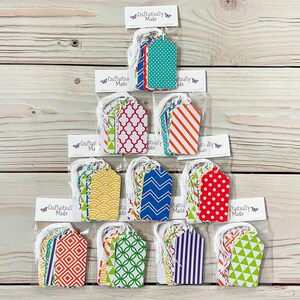 May include: A set of 12 gift tags featuring a variety of colourful geometric patterns. The tags are packaged in clear plastic bags with white twine and a "Craftastailly Made" label.
