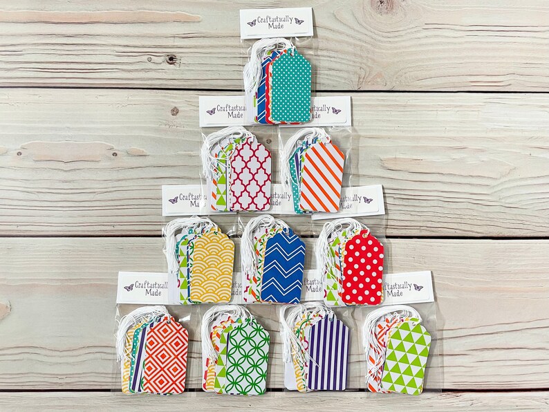 May include: A set of 12 gift tags featuring a variety of colourful geometric patterns. The tags are packaged in clear plastic bags with white twine and a "Craftastailly Made" label.
