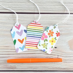 May include: Three decorative gift tags with white string, featuring colorful patterns: hearts, rainbow stripes, and flowers. An orange pen rests below the tags on a light wood surface.