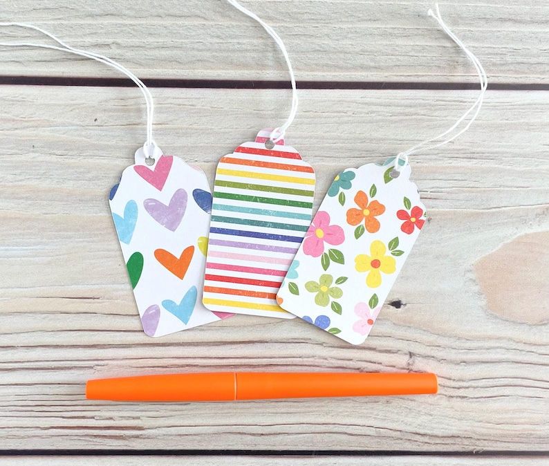 May include: Three decorative gift tags with white string, featuring colorful patterns: hearts, rainbow stripes, and flowers. An orange pen rests below the tags on a light wood surface.