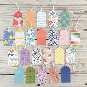 May include: A collection of colorful gift tags with various patterns, including floral, polka dots, and stripes. Some tags have text, and all have white string attached. The tags are arranged on a light wood surface.
