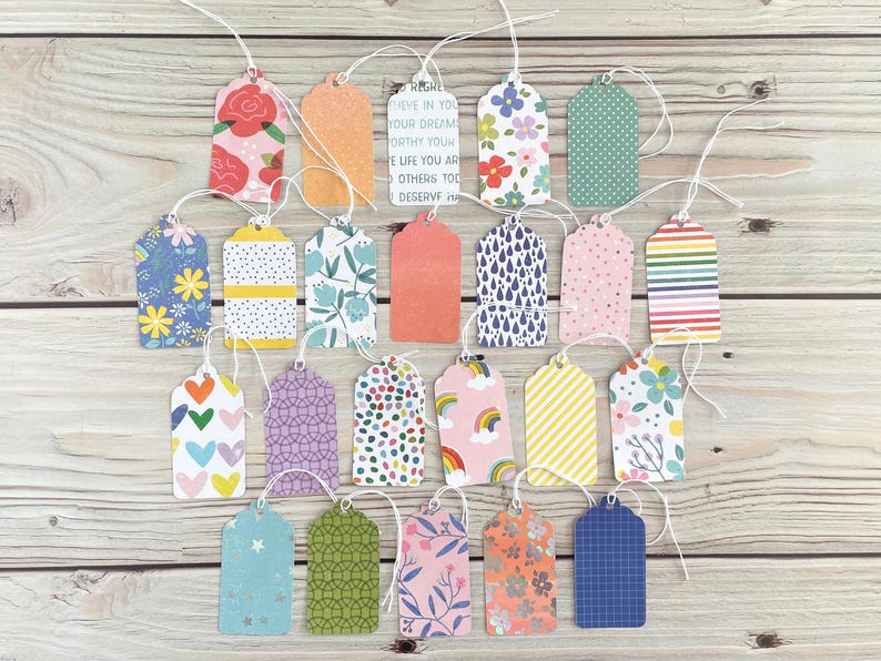 May include: A collection of colorful gift tags with various patterns, including floral, polka dots, and stripes. Some tags have text, and all have white string attached. The tags are arranged on a light wood surface.