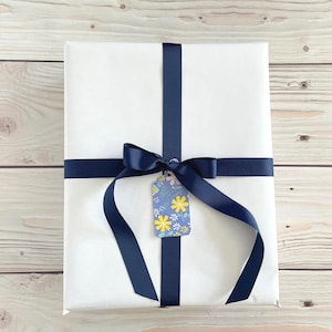 May include: A wrapped gift with white paper, navy blue ribbon, and a floral gift tag. The ribbon is tied in a bow and crosses the package. The gift tag has a blue background with yellow and white flowers.