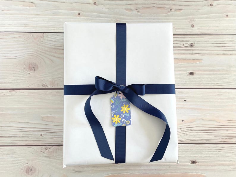 May include: A wrapped gift with white paper, navy blue ribbon, and a floral gift tag. The ribbon is tied in a bow and crosses the package. The gift tag has a blue background with yellow and white flowers.