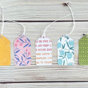 May include: Five decorative gift tags with white string. The tags feature various patterns, including yellow polka dots, pink floral, blue floral, green geometric, and a tag with text that reads "ELF BE FREE OF E JOY KEEP C FE NEVER GIVE AND GROW Y OMPLISH ANY ND TO YOURS."
