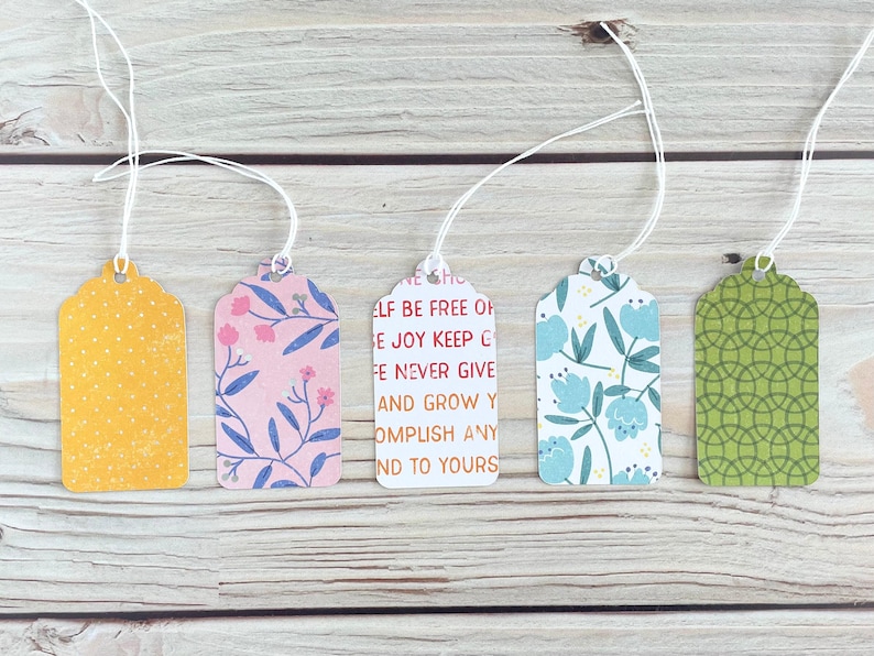May include: Five decorative gift tags with white string. The tags feature various patterns, including yellow polka dots, pink floral, blue floral, green geometric, and a tag with text that reads "ELF BE FREE OF E JOY KEEP C FE NEVER GIVE AND GROW Y OMPLISH ANY ND TO YOURS."