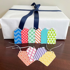 May include: A set of eight colourful gift tags with geometric patterns. The tags are made of paper and have a string attached to them. The tags are arranged in two rows on a wooden surface. The tags are perfect for adding a personal touch to gifts.
