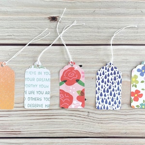 May include: Five decorative gift tags with white string. The tags feature various designs, including an orange gradient with white dots, text, red roses on pink, blue raindrops, and colorful flowers on white. They are arranged on a wooden surface.