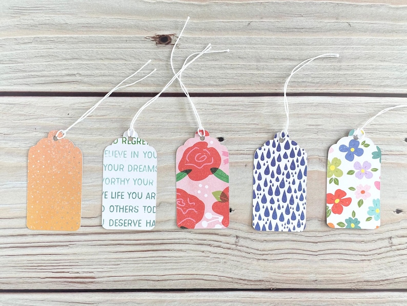 May include: Five decorative gift tags with white string. The tags feature various designs, including an orange gradient with white dots, text, red roses on pink, blue raindrops, and colorful flowers on white. They are arranged on a wooden surface.