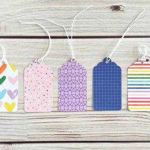 May include: Five decorative gift tags with white string. The tags feature various patterns: colorful hearts, pink polka dots, a purple geometric design, a blue grid, and rainbow stripes. They are arranged on a light wood surface.