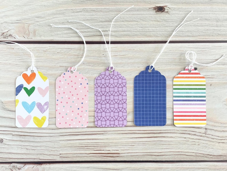 May include: Five decorative gift tags with white string. The tags feature various patterns: colorful hearts, pink polka dots, a purple geometric design, a blue grid, and rainbow stripes. They are arranged on a light wood surface.
