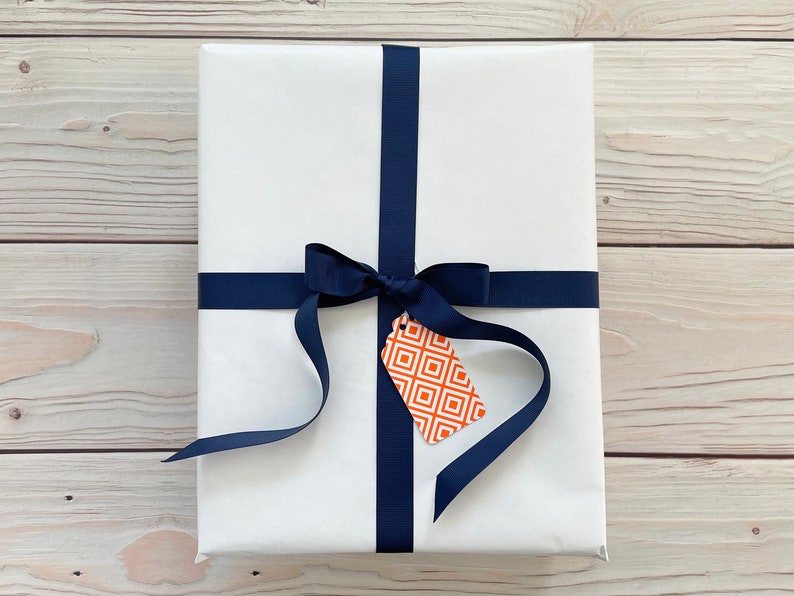 May include: A white gift box with a navy blue ribbon and bow. A small orange and white gift tag with a geometric pattern is attached to the ribbon.