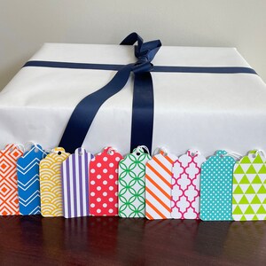 May include: A white gift box with a navy blue ribbon and eight colourful gift tags with geometric patterns. The tags have different patterns, including stripes, polka dots, and geometric shapes.