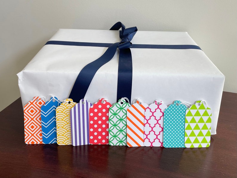 May include: A white gift box with a navy blue ribbon and eight colourful gift tags with geometric patterns. The tags have different patterns, including stripes, polka dots, and geometric shapes.