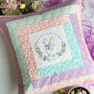 Könnte beinhalten: Ein quadratisches Kissen mit einem pastellrosa, grünen und violetten Patchwork-Muster. In der Mitte des Kissens befindet sich ein gestickter Hase mit einem Blumenkranz. Das Kissen ist von Ostereiern und Blumen umgeben.