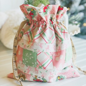 May include: A pink and green patchwork fabric gift bag with a drawstring closure. The bag features a reindeer print and a plaid pattern. The bag is tied with a brown ribbon.