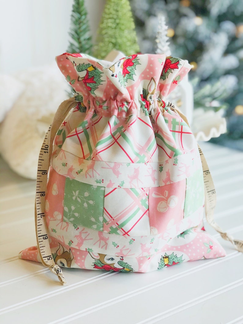 May include: A pink and green patchwork fabric gift bag with a drawstring closure. The bag features a reindeer print and a plaid pattern. The bag is tied with a brown ribbon.