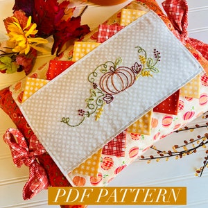 May include: A PDF pattern for a pillow with a stitched pumpkin design. The pillow is made of various fabrics in shades of orange, red, and yellow. The pillow has a bow made of red and white checkered fabric. The text "PDF PATTERN" is displayed in a yellow banner.