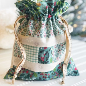 May include: A green and white fabric gift bag with a drawstring closure. The bag is decorated with a variety of holiday-themed patterns, including snowflakes, holly, and plaid. The bag is tied with a white ribbon.