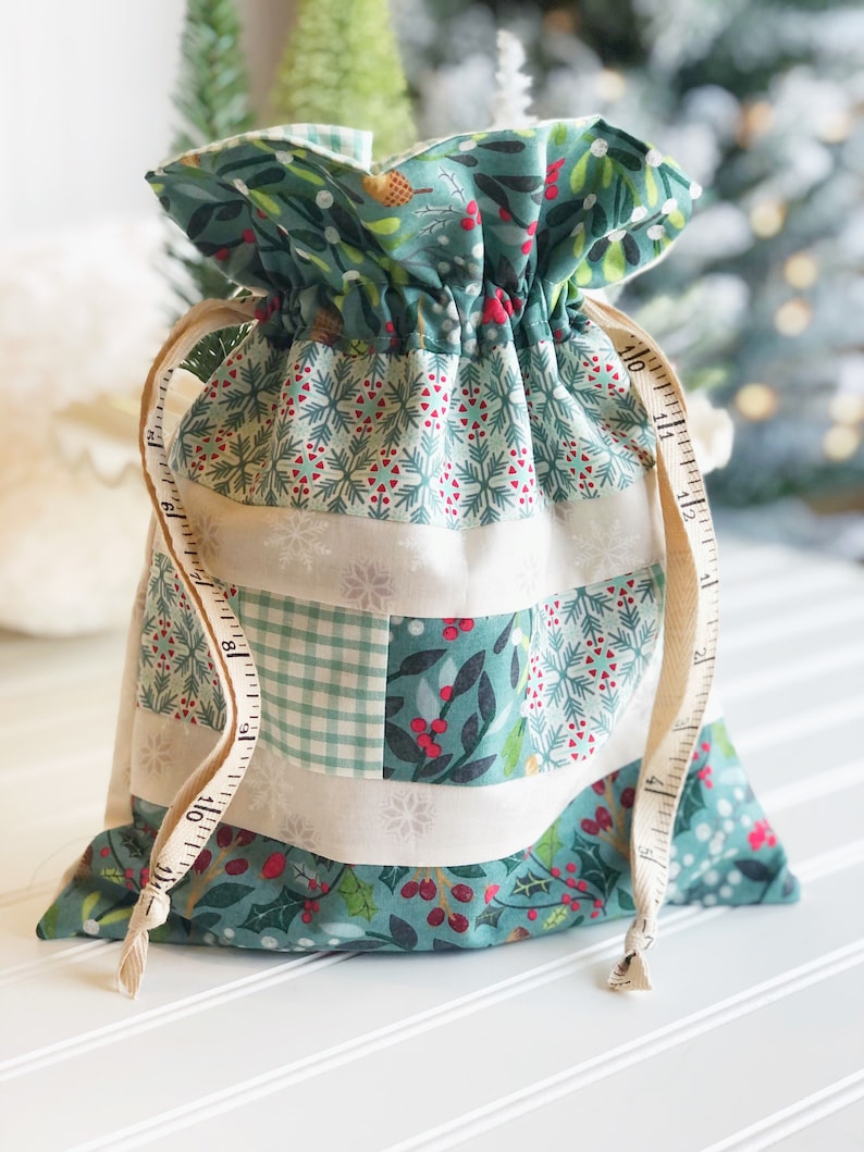 May include: A green and white fabric gift bag with a drawstring closure. The bag is decorated with a variety of holiday-themed patterns, including snowflakes, holly, and plaid. The bag is tied with a white ribbon.